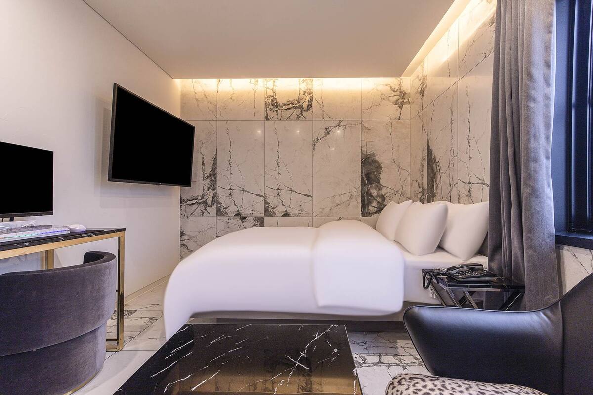 Una habitación de hotel moderna con una cama grande, un escritorio con computadora, un televisor y una zona de estar con una silla y un sofá.