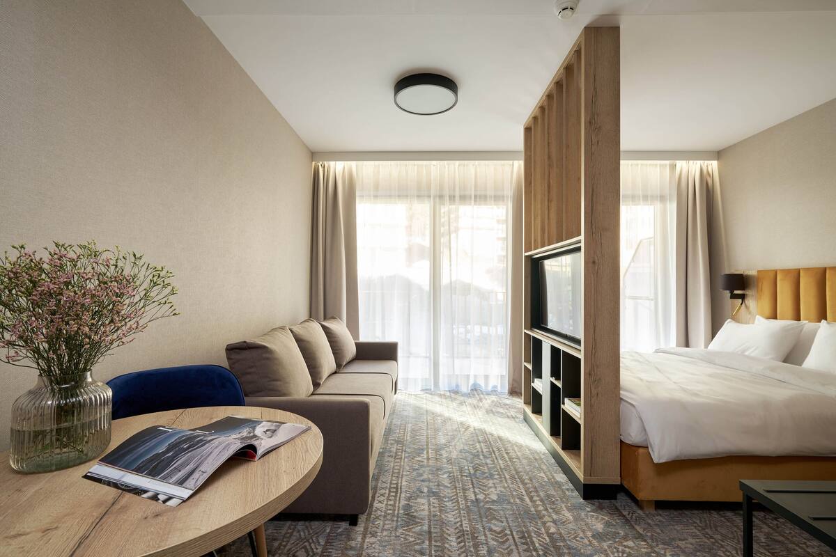 A hotel room with a sofa, a bed, a TV, a table with a magazine, and a vase with flowers.