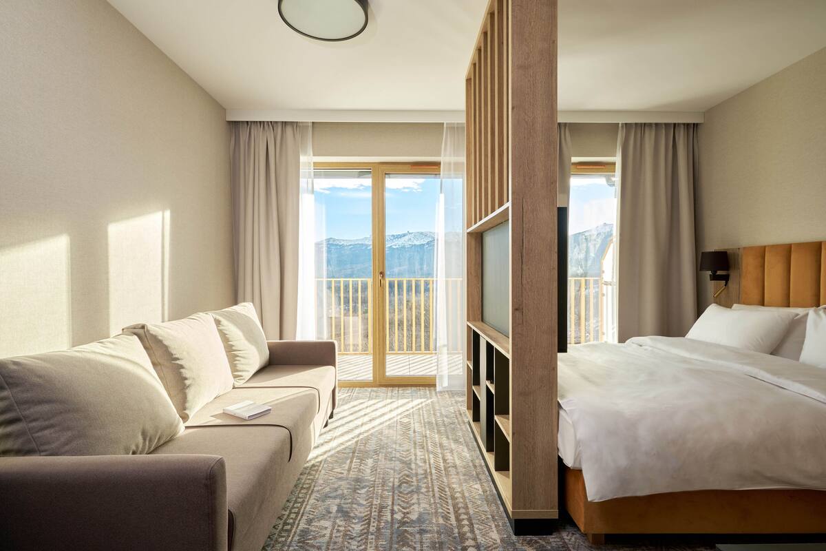 A hotel room with a bed, a sofa, a TV, and a balcony with a mountain view.