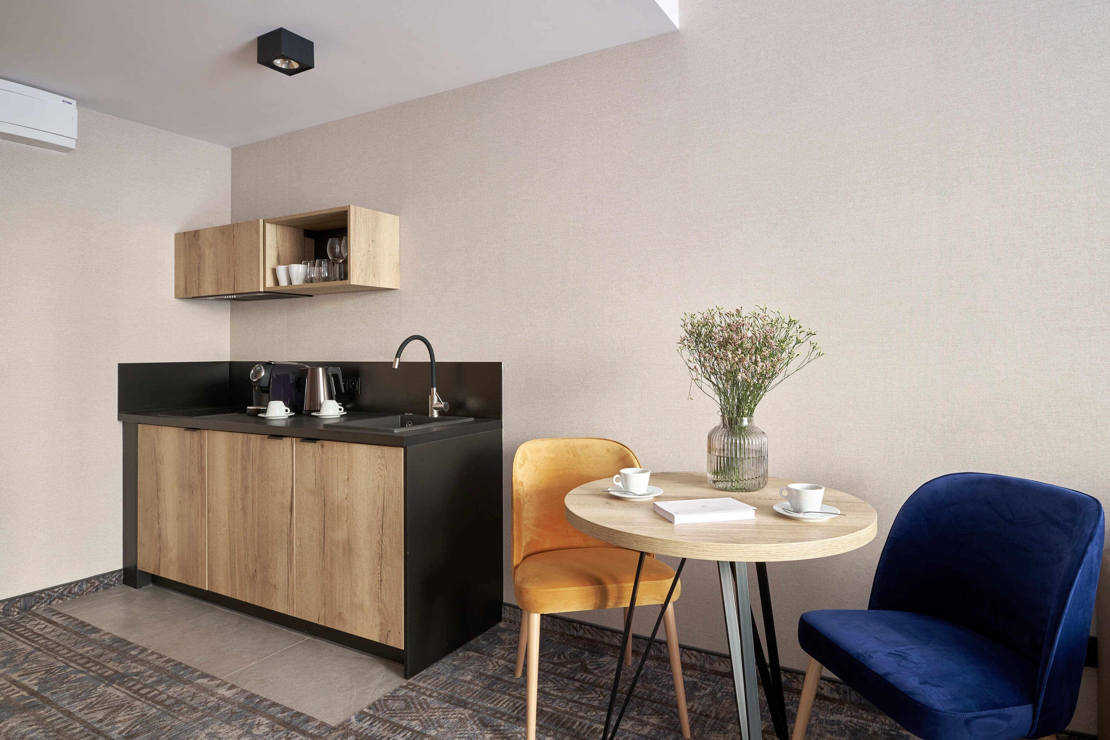 A compact kitchen with a black countertop, wooden cabinets, and a built-in sink. There is a small round table with two chairs, one yellow and one blue, and a vase with flowers on the table.