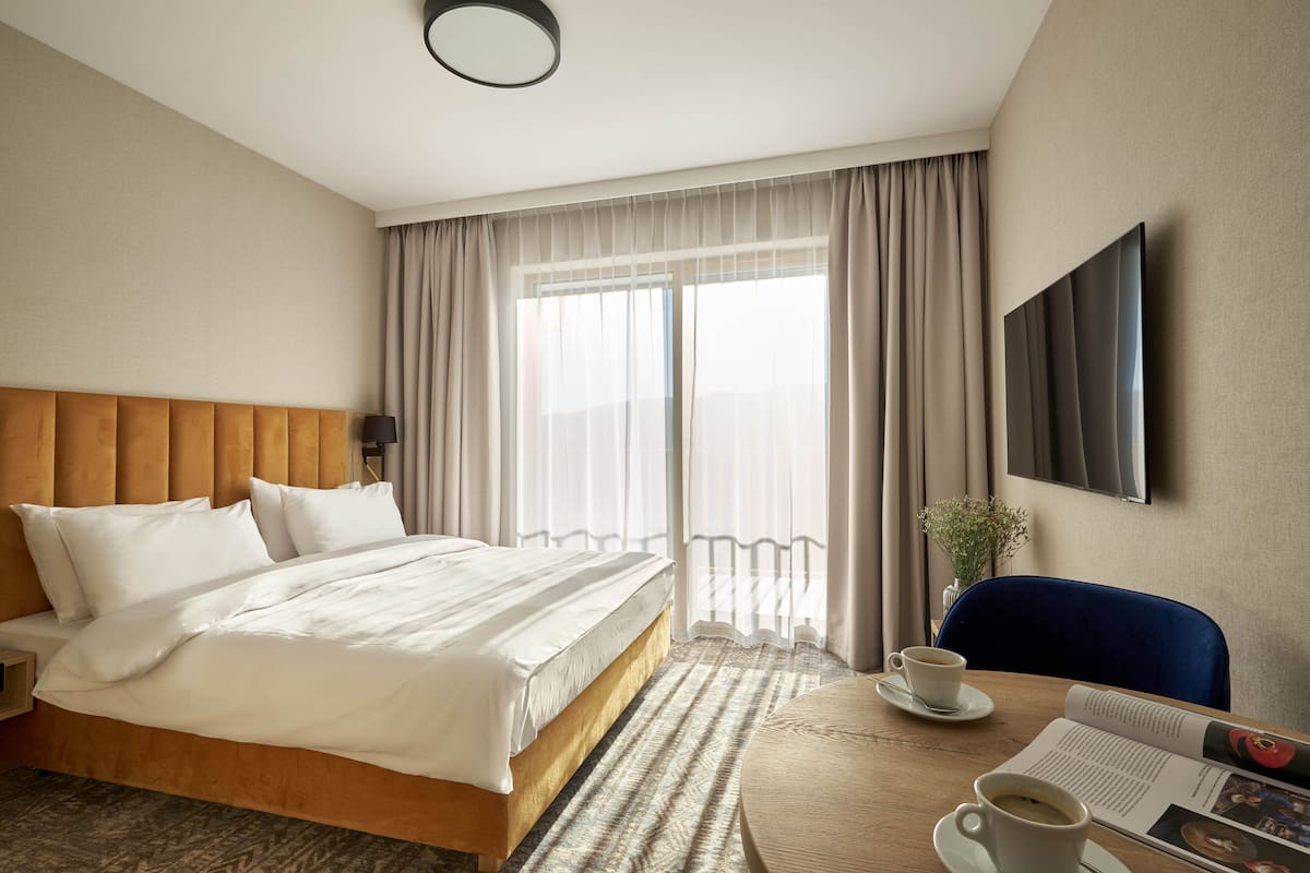 A hotel room with a large bed, a round table with a cup and saucer, a chair, a TV mounted on the wall, and a window with curtains.