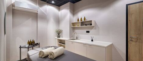 A modern spa room with a massage table, a small sink, and shelves with bottles.