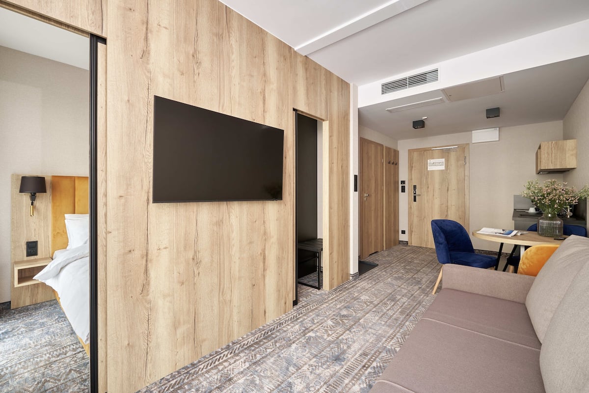 A hotel room with a wooden wall, a flat-screen TV, a sofa, a dining table with a vase, and a blue chair.