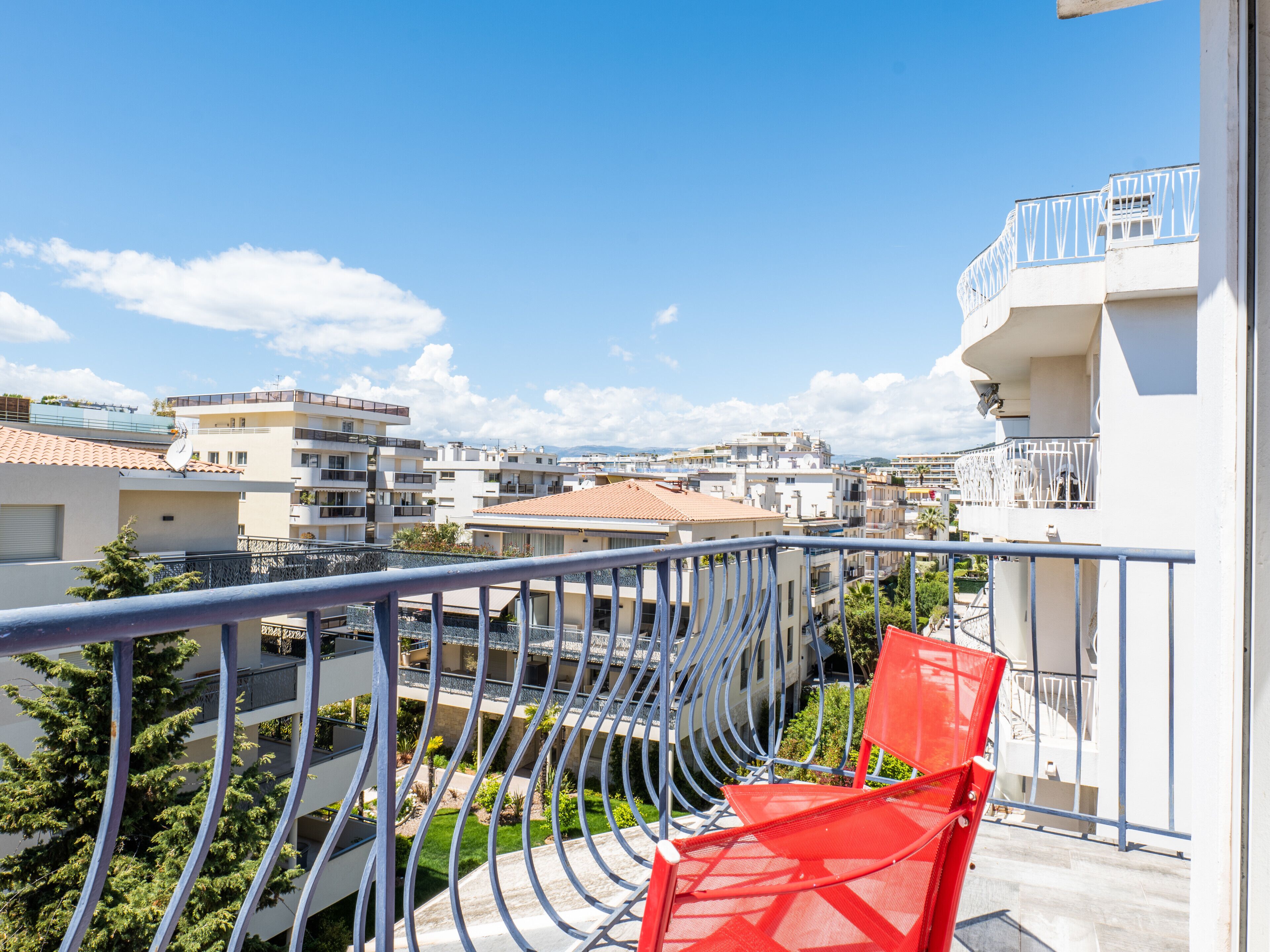 A balcony with a red chair, overlooking a residential area with multiple buildings and a clear blue sky.