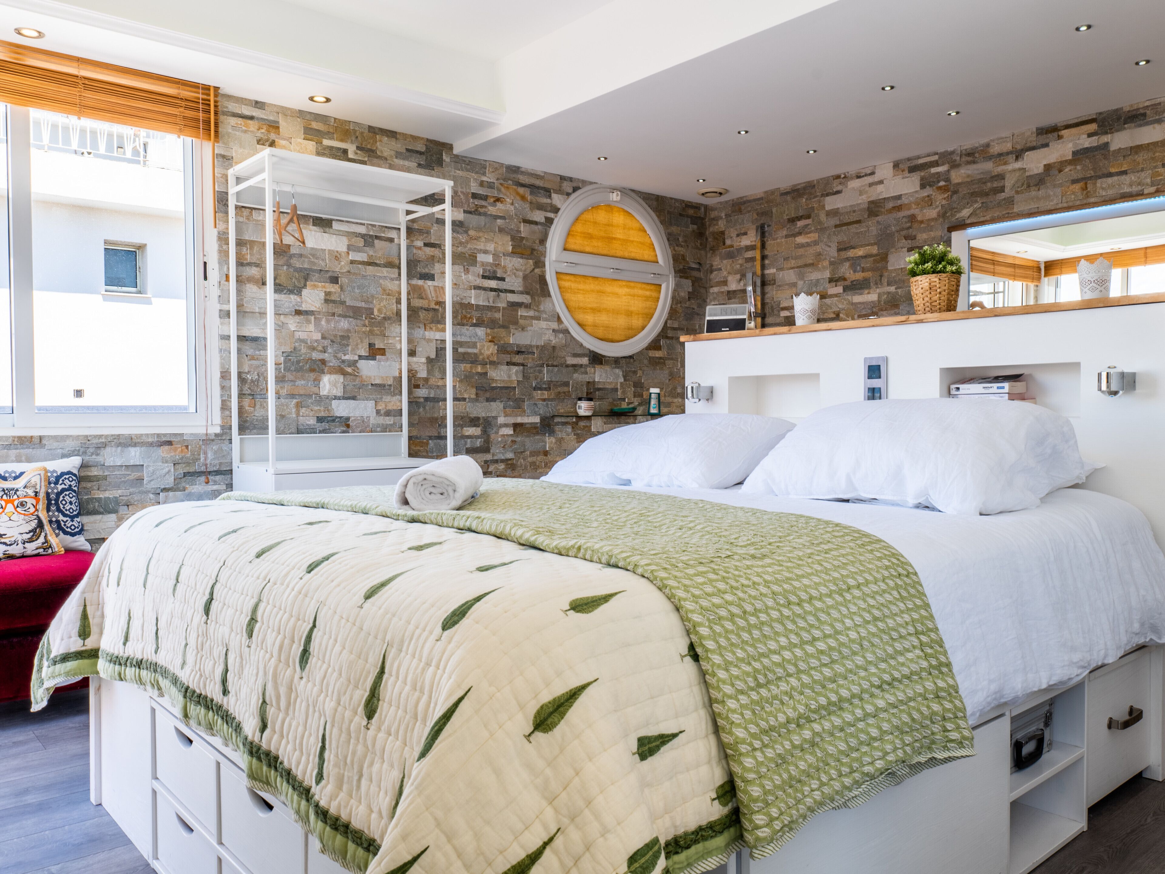 A modern bedroom with a stone wall, a bed with a green leaf-patterned blanket, a white bedside table, and a large mirror above the bed.
