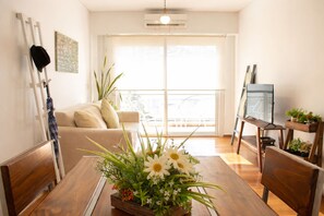 A living room with a wooden dining table, a sofa, a TV on a stand, and a balcony with a railing.