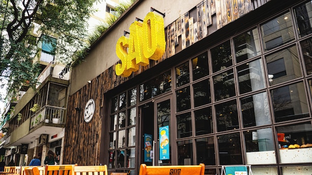 Un edificio con ampie vetrate, posti a sedere all'aperto con sedie e tavoli di legno, e un cartello giallo ben visibile.