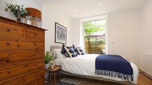 A bedroom with a wooden dresser, a bed with blue and white bedding, a window with a view of greenery, and a framed picture on the wall. - Archway luxury interior designed 1 bed (London)