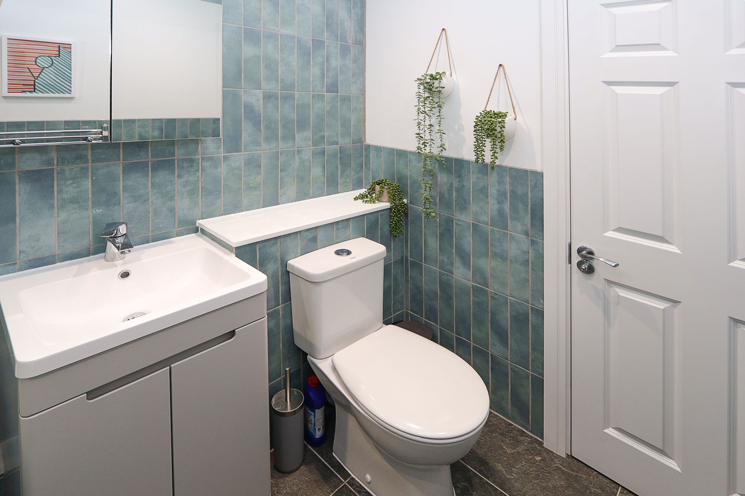 Un bagno con lavandino, water e porta bianchi. Le pareti sono rivestite di piastrelle blu e c'è una piccola mensola con piante.