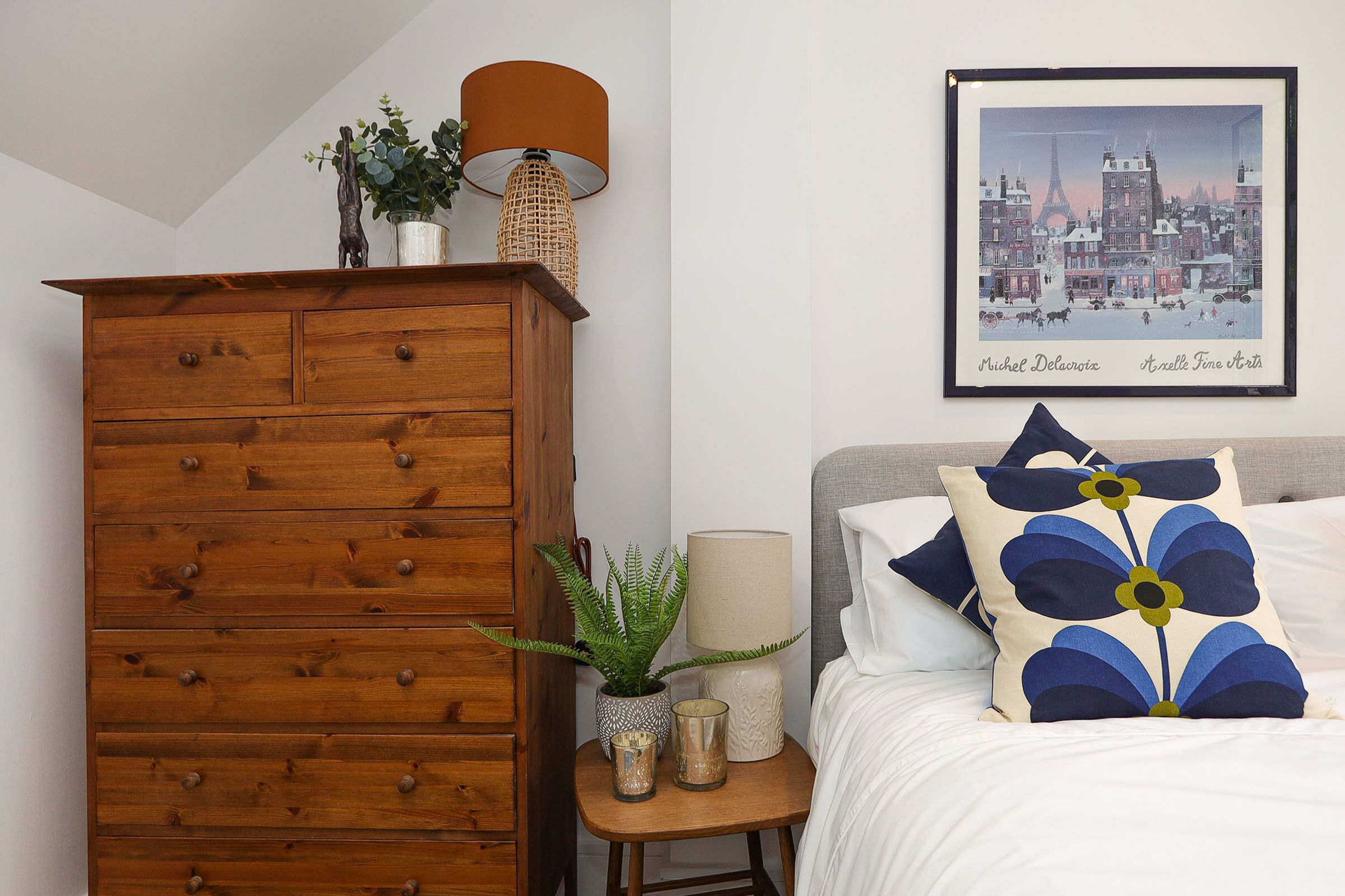 Een slaapkamer met een houten dressoir, een nachtkastje met een lamp, een lijst met een stadsgezicht en een bed met blauwe en witte bloemenkussens.