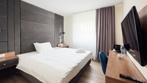 Quarto de hotel com cama, escrivaninha, cadeira, televisão e janela com cortinas.