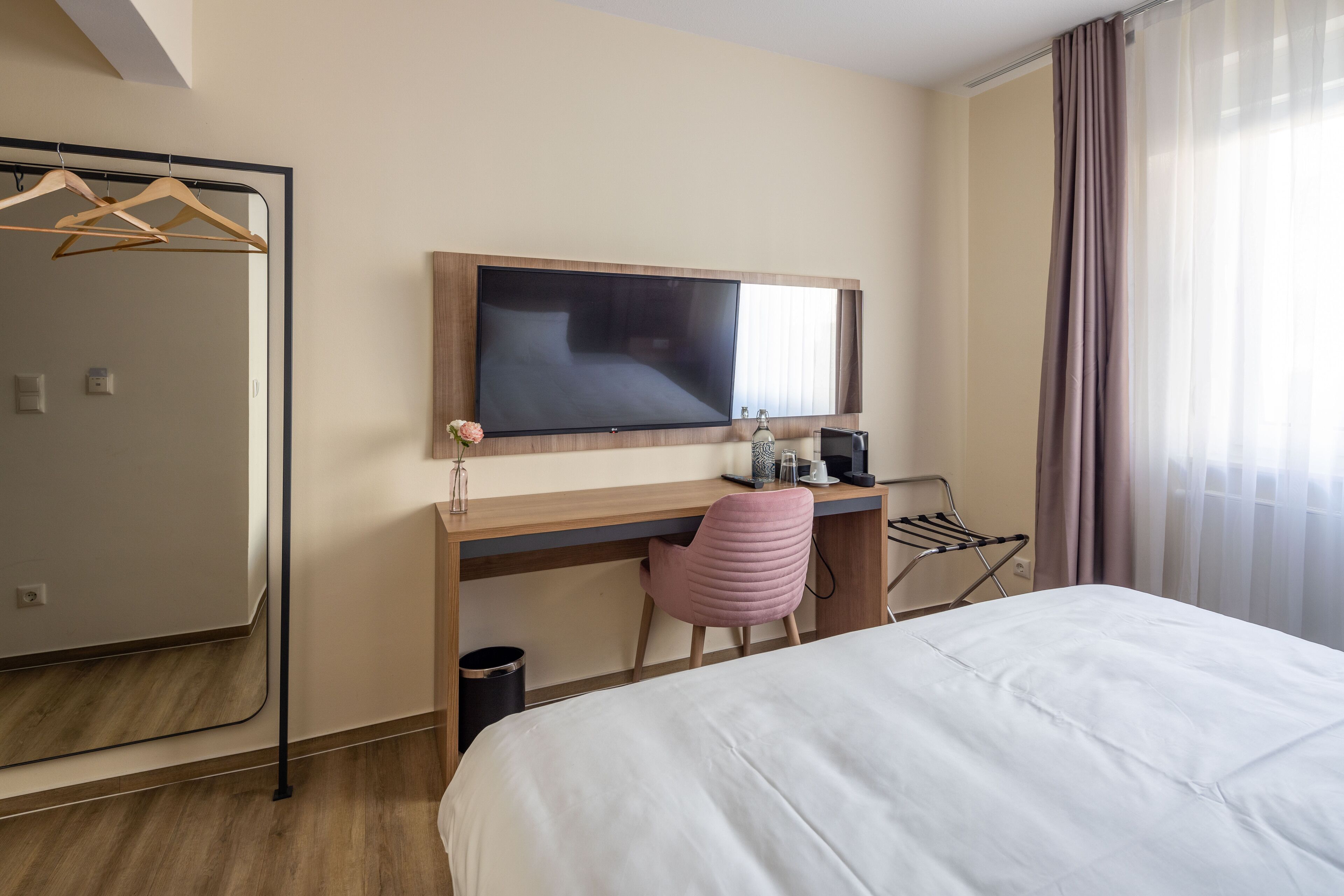 A hotel room with a bed, a desk with a chair, a television, a mirror, and a window with curtains.