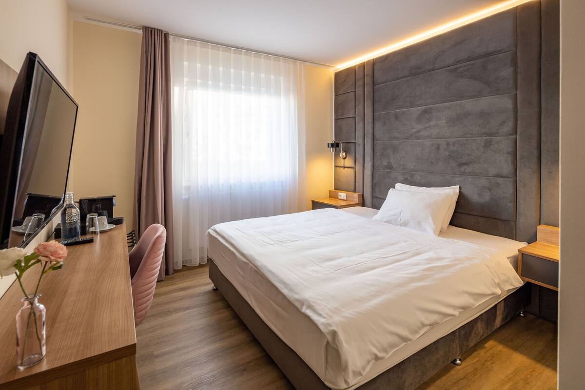 Hotelkamer met een groot bed, een televisie, een bureau met een bloemetjesvaas en een raam met gordijnen.