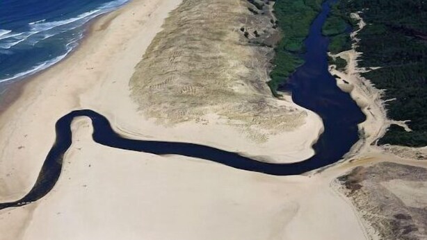 Luchtbeeld van een kustgebied met een lang zandstrand, een rivier die parallel aan de kust loopt, en dichtbegroeide groene bosgebieden.