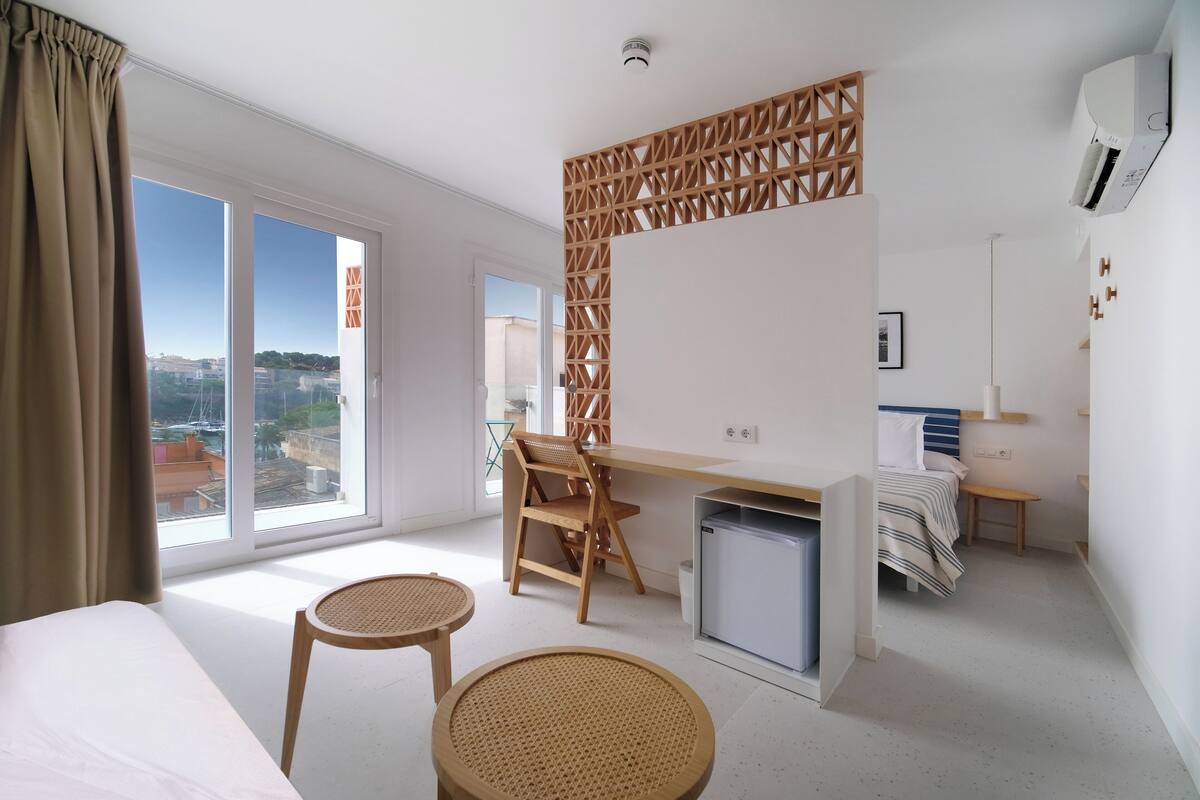 A modern hotel room with a large window, a desk, a chair, a bed, and a small kitchenette.