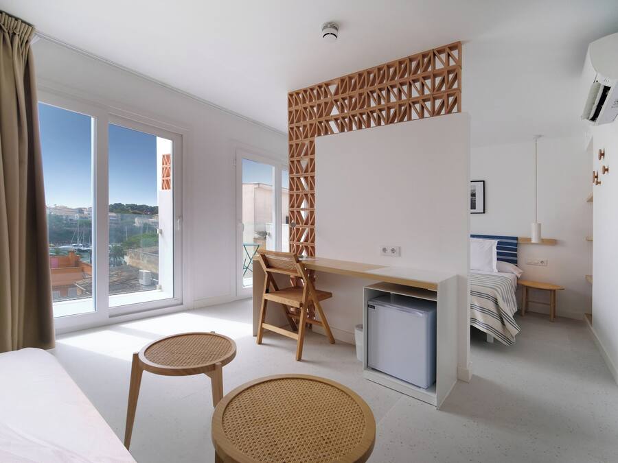 A modern hotel room with a large window, a desk, a chair, a bed, and a small kitchenette.