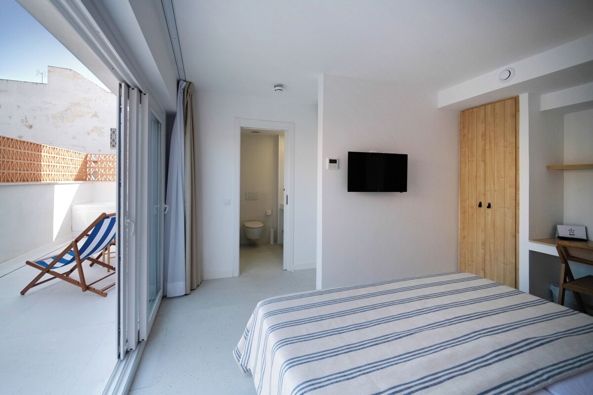 A bedroom with a bed, a TV, a wooden wardrobe, and a balcony with a chair.
