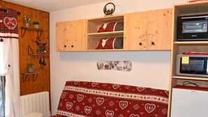A cozy room with a red patterned sofa, wooden shelves with decorative items, a white radiator, and a small white stool. - 22m² Studio in Saint-Jean-d'Aulps, near slopes (Saint-Jean-d'Aulps)