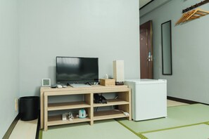 A room with a TV on a wooden stand, a small white fridge, a trash bin, and a door with a mirror and clothes hangers.
