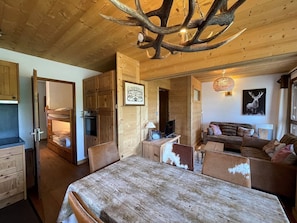 Interior rústico de cabana com mesa de jantar de madeira, sofá, televisão e lareira.
