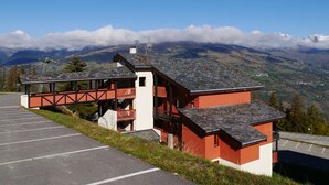 Ein rot-weißes Gebäude mit Balkon, Parkplatz und einer Berglandschaft im Hintergrund.