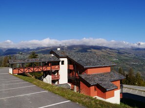Ein rot-weißes Gebäude mit Balkon, Parkplatz und einer Berglandschaft im Hintergrund.