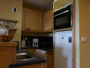 Una cocina compacta con armarios de madera, microondas, refrigerador, cafetera y un lavamanos.