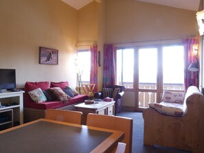 Una sala de estar acogedora con un sofá, una mesa de comedor y un televisor. Hay ventanas amplias con cortinas, una lámpara y un cuadro en la pared.
