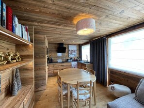 Interior acolhedor de uma cabana, com uma mesa de jantar de madeira, cadeiras, um sofá e uma estante com livros e objetos decorativos.