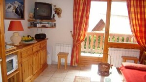 Ein gemütliches Wohnzimmer mit Fernseher, Holzschränken, einem kleinen Tisch und einem Balkon mit Vorhängen.