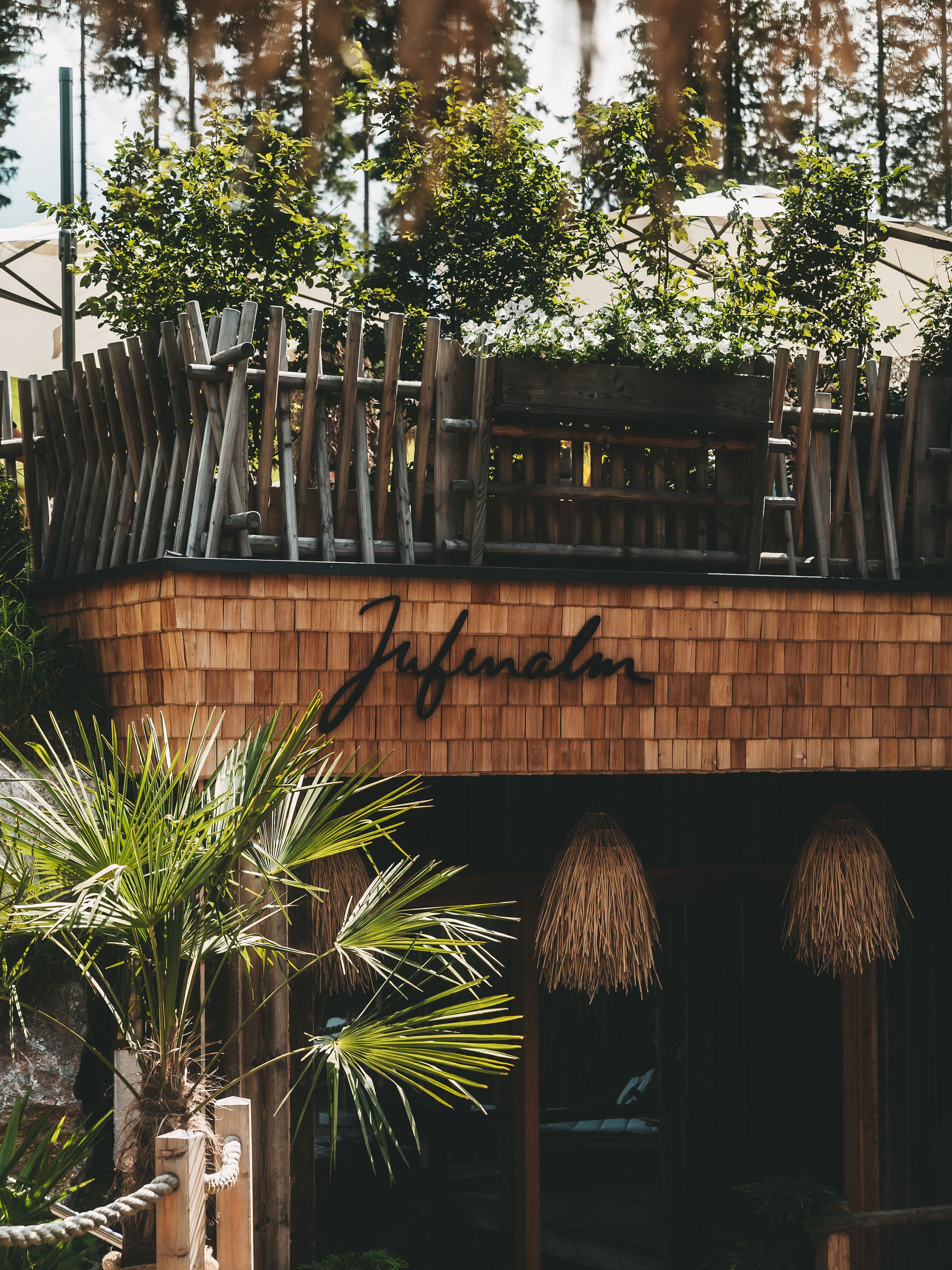 A wooden structure with the name 'Jufalmi' carved into it, surrounded by greenery and a rope barrier.