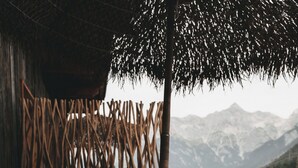 Une terrasse en bois offrant une vue sur les montagnes, une clôture en bambou et un espace détente avec des coussins et un vase.
