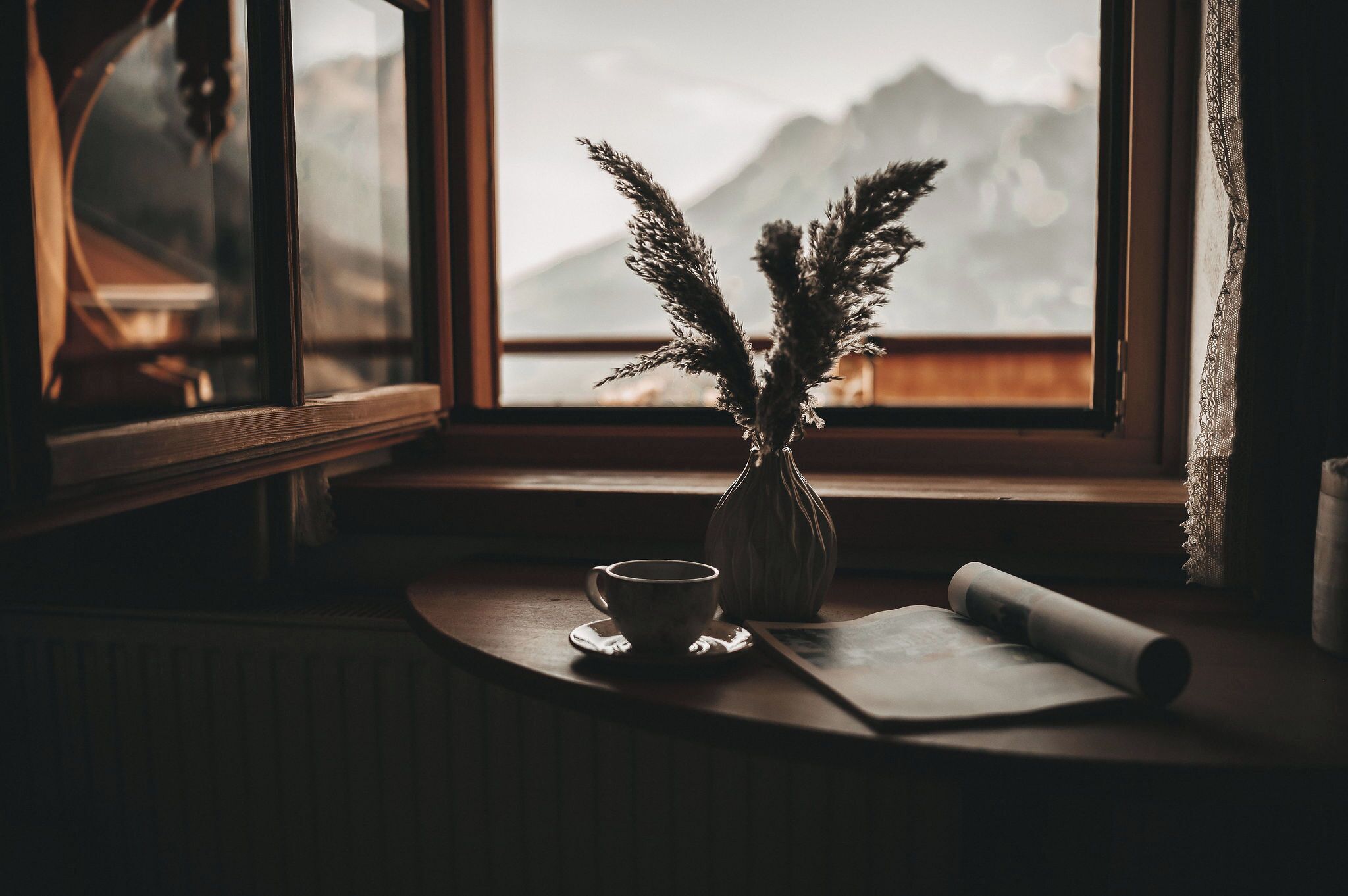 A window with a view of mountains, a vase with dried plants, a cup, and a rolled-up paper on a wooden table.