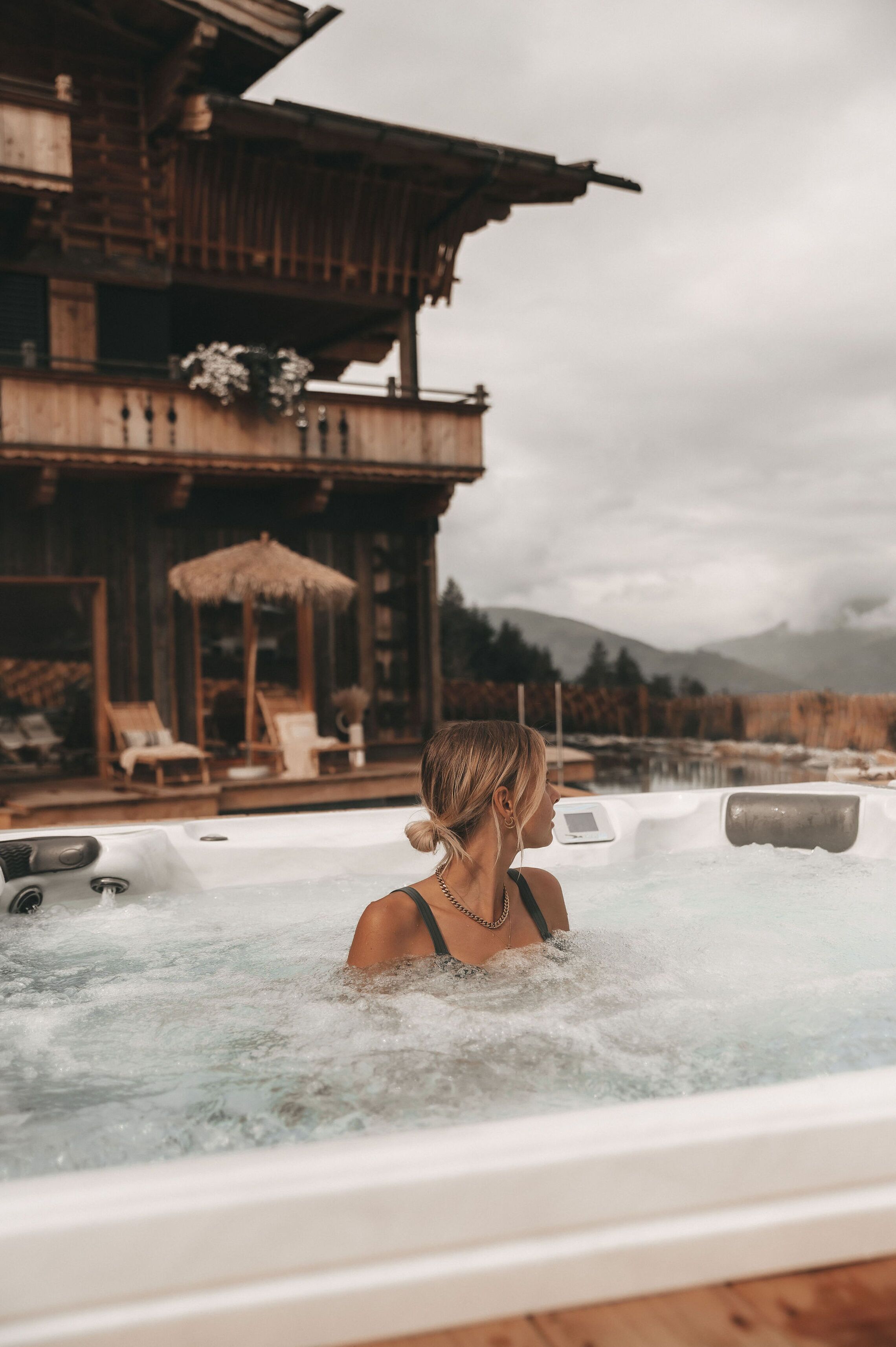 Eine Person in einem Whirlpool vor einem Holzgebäude mit Liegestühlen im Hintergrund.