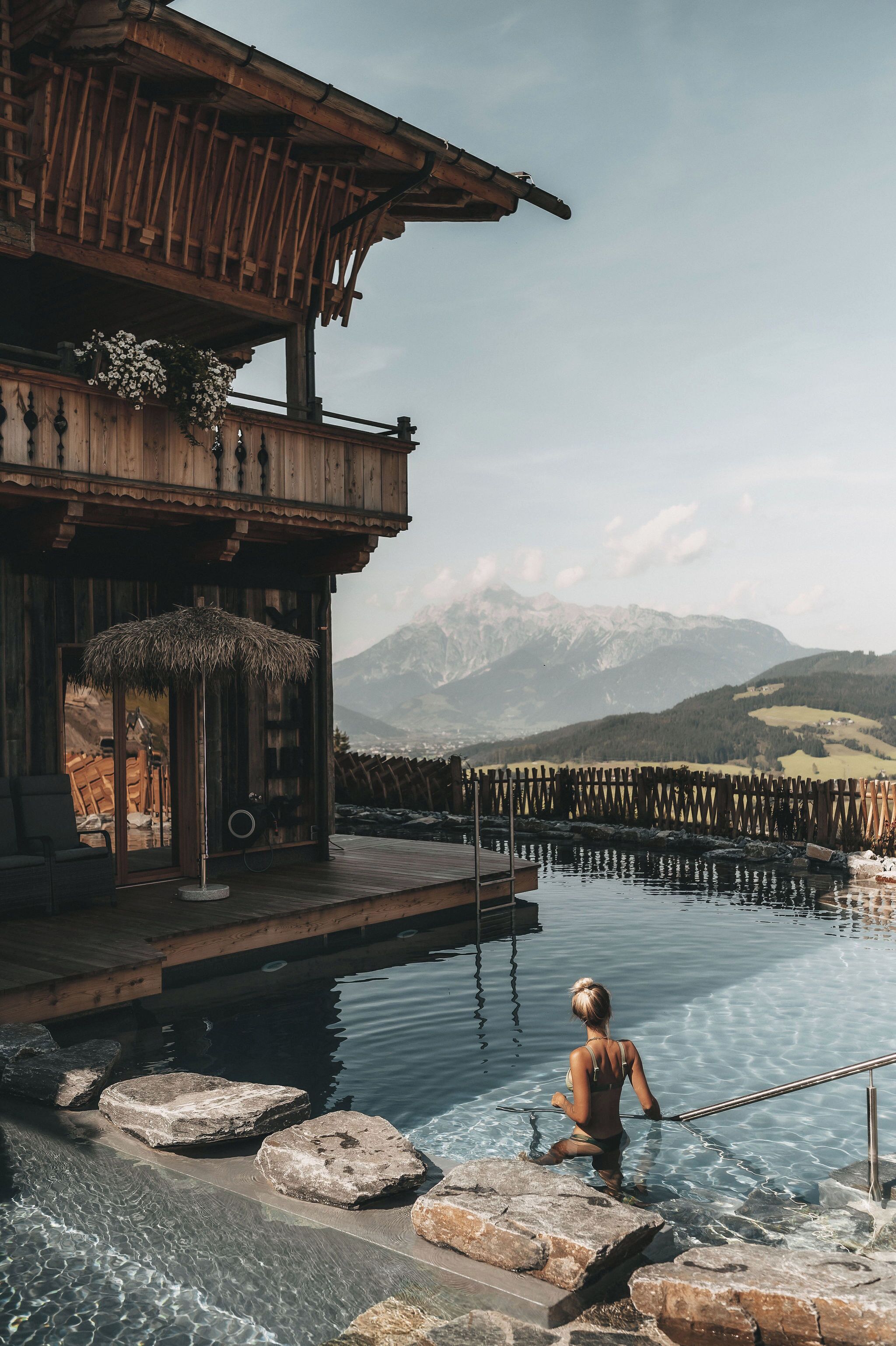 Un bâtiment en bois doté d’un balcon donnant sur une piscine bordée de rochers et offrant une vue sur la montagne.