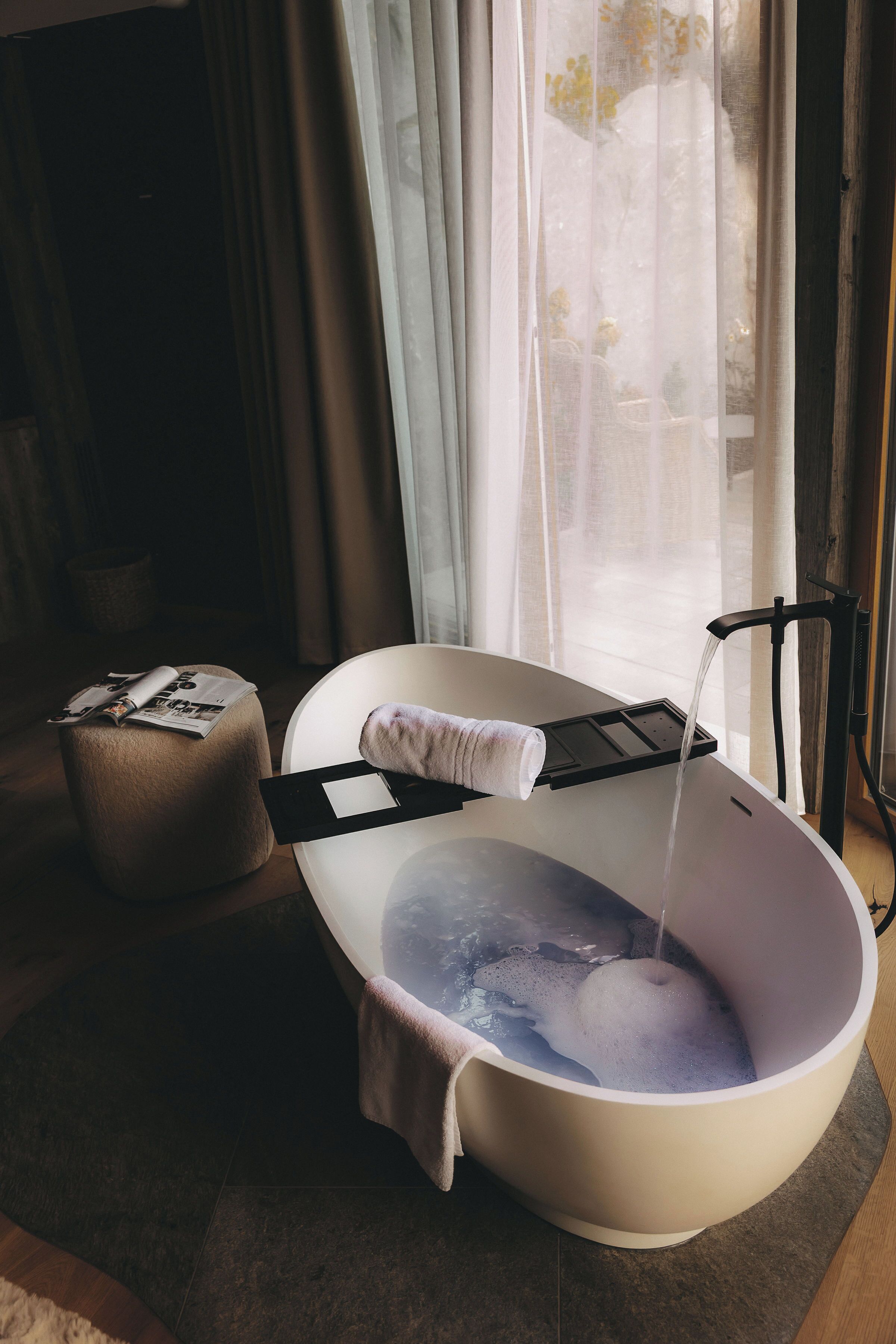 A modern bathroom with a freestanding bathtub, a towel rack, and a small stool with a magazine.