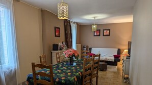 A cozy living room with a dining area, featuring a wooden dining table with chairs, a sofa, a TV, and a fireplace. - Charming Cottage in Tranquil Village with Private Parking and Baby Equipment (Villemurlin)