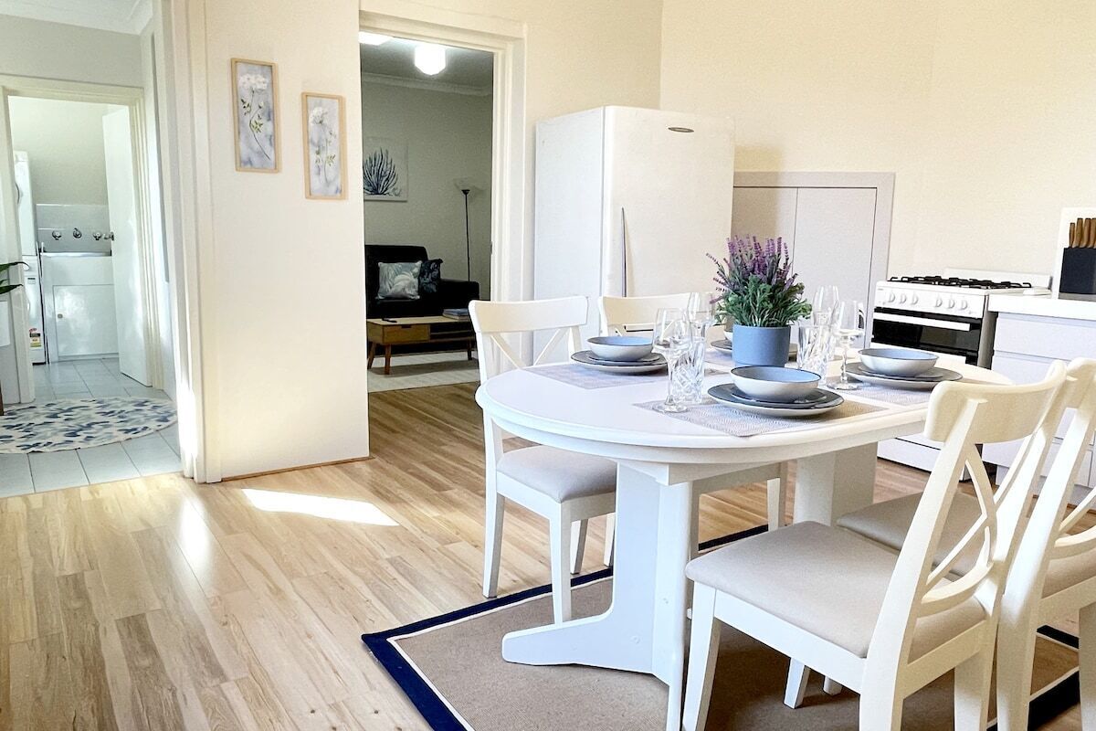Uma cozinha moderna com eletrodomésticos brancos, uma mesa de jantar posta para quatro pessoas e vista para a sala de estar.