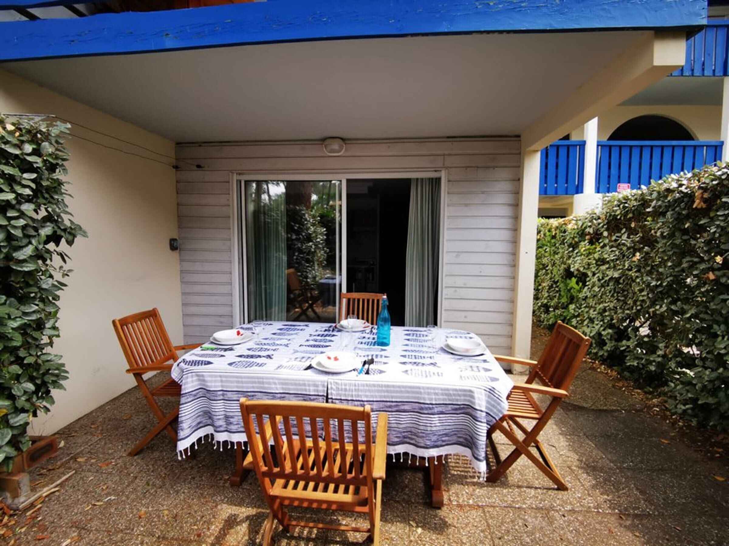 Eine Terrasse im Freien mit einem Tisch für zwei Personen, umgeben von Holzstühlen, und einer Glastür, die zu einem überdachten Bereich führt.