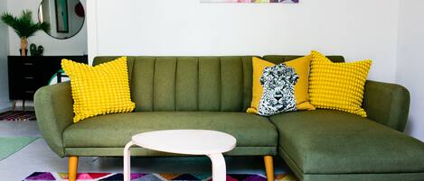 Un canapé d’angle vert avec des coussins à motifs jaunes et noirs, une table basse blanche et un tapis géométrique aux couleurs vives.