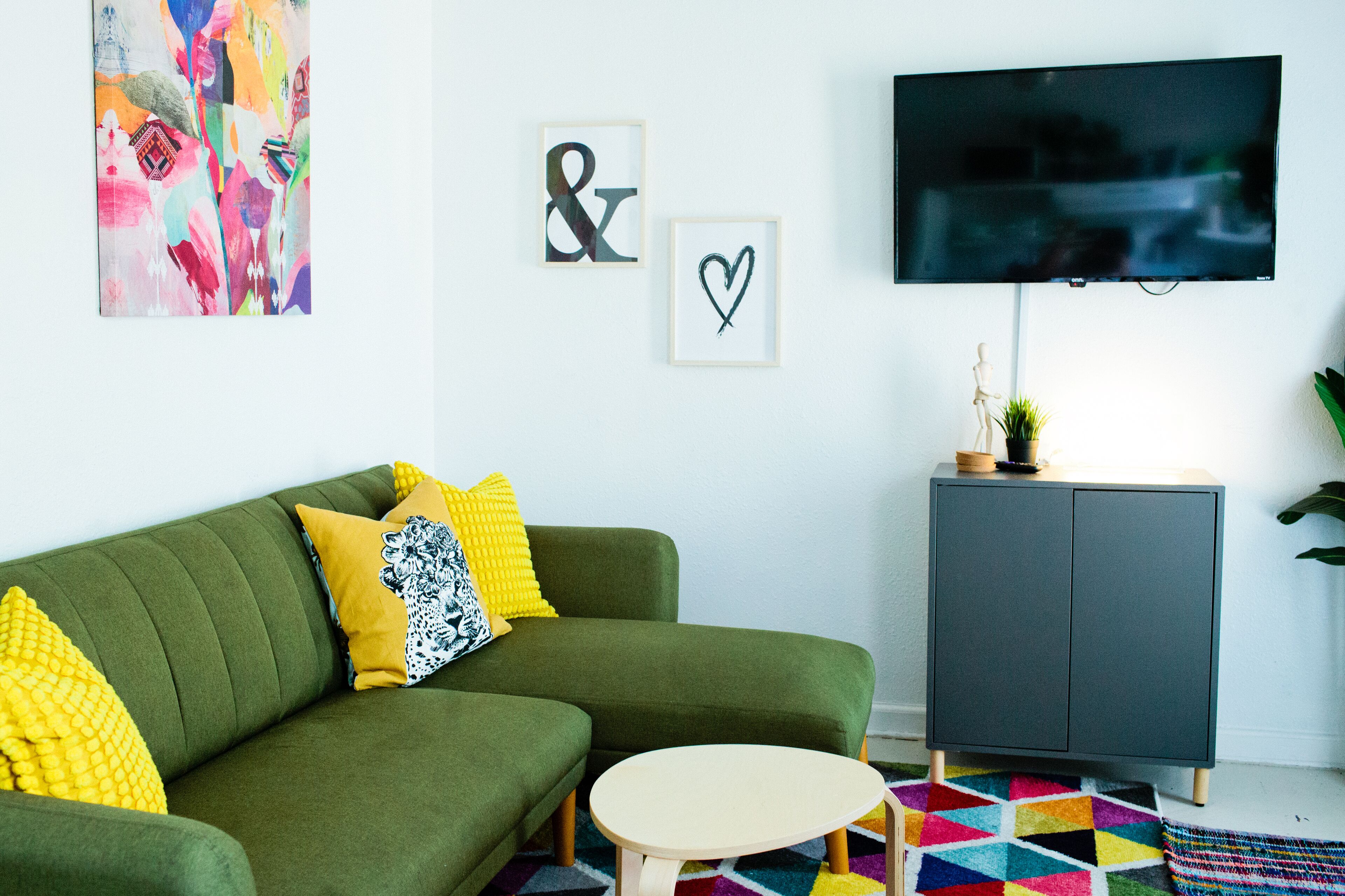 Een moderne woonkamer met een groene bank, gele en zwarte kussens, een klein rond tafeltje, een grijze kast en een kleurrijk vloerkleed.