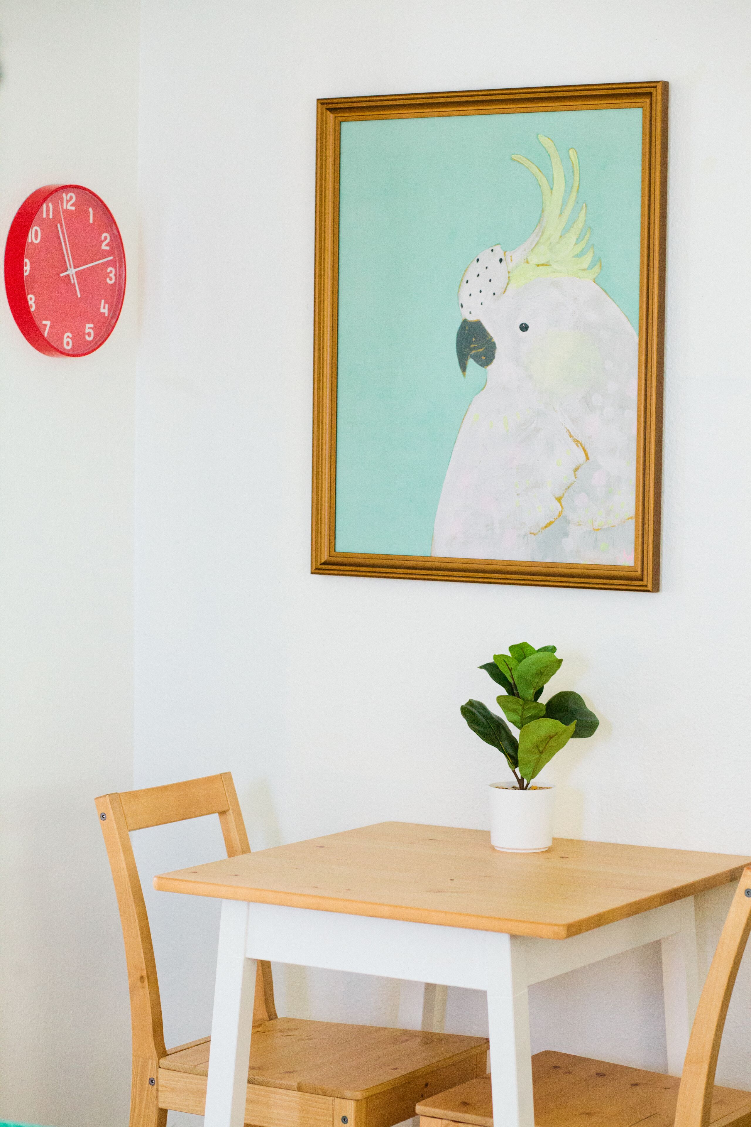 A room with a wooden table, two chairs, a red clock, a framed painting of a bird, and a potted plant.