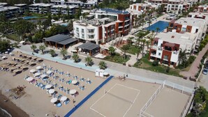 An aerial view of a coastal hotel area with multiple buildings, a beach with sun loungers and umbrellas, and a tennis court. - SUNDANCE SUITES HOTEL (Bodrum)