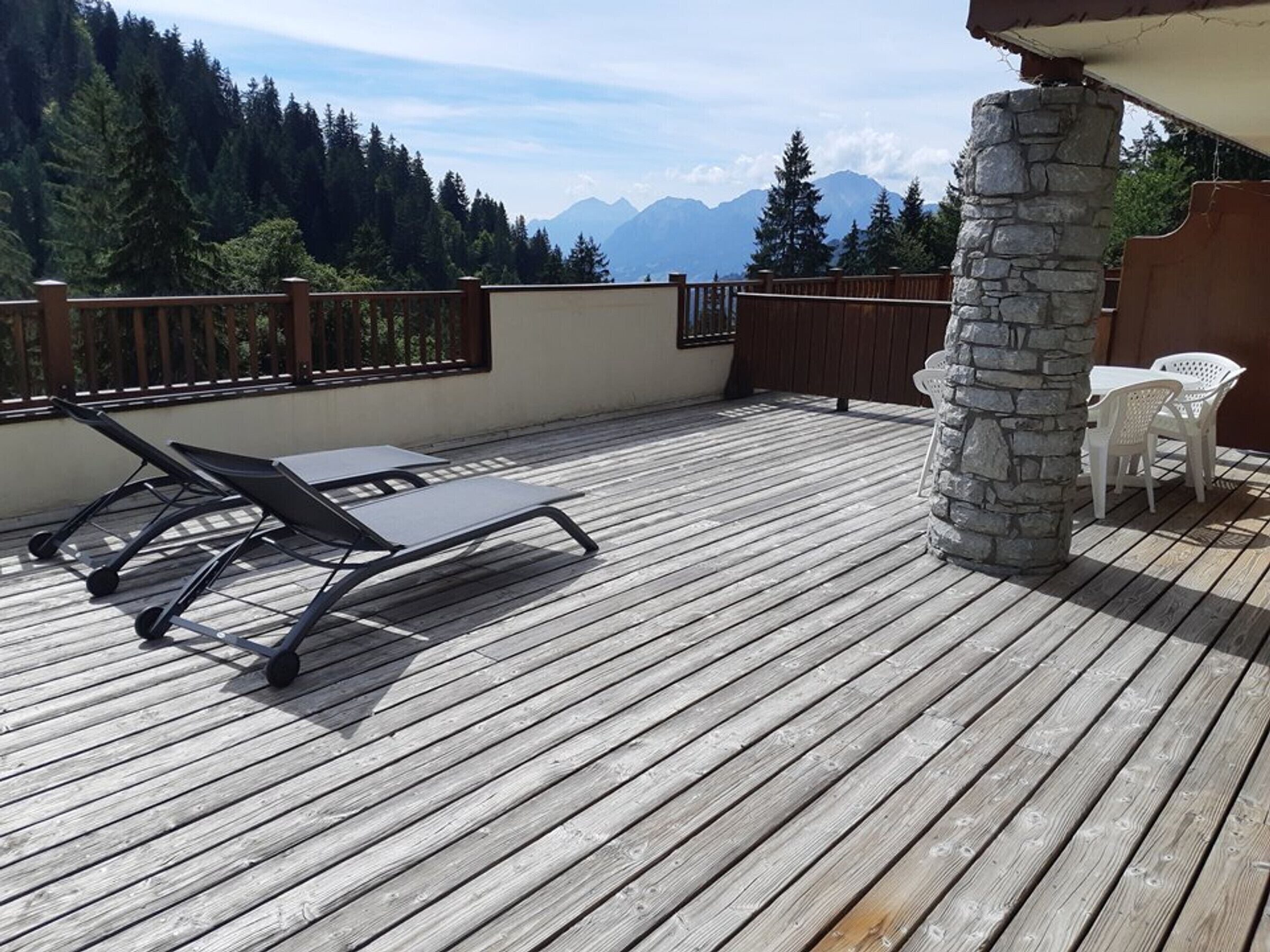 A wooden deck with a lounge chair and a table, overlooking a forest and mountains.