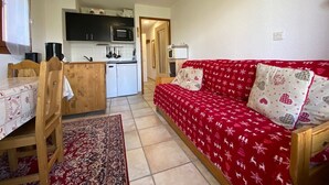 En kompakt opholdsstue med tekøkken, spiseplads med bord og stole, samt en rød sofa med mønstrede puder.