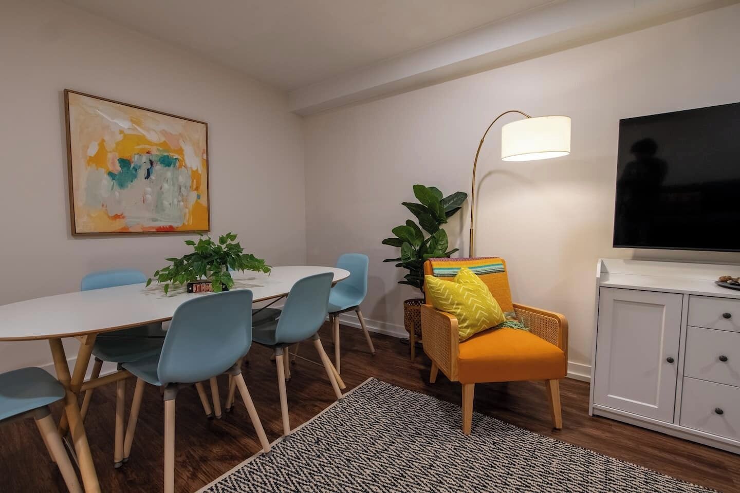 A dining area with a round table, blue chairs, a yellow cushioned chair, a floor lamp, and a TV on a cabinet.