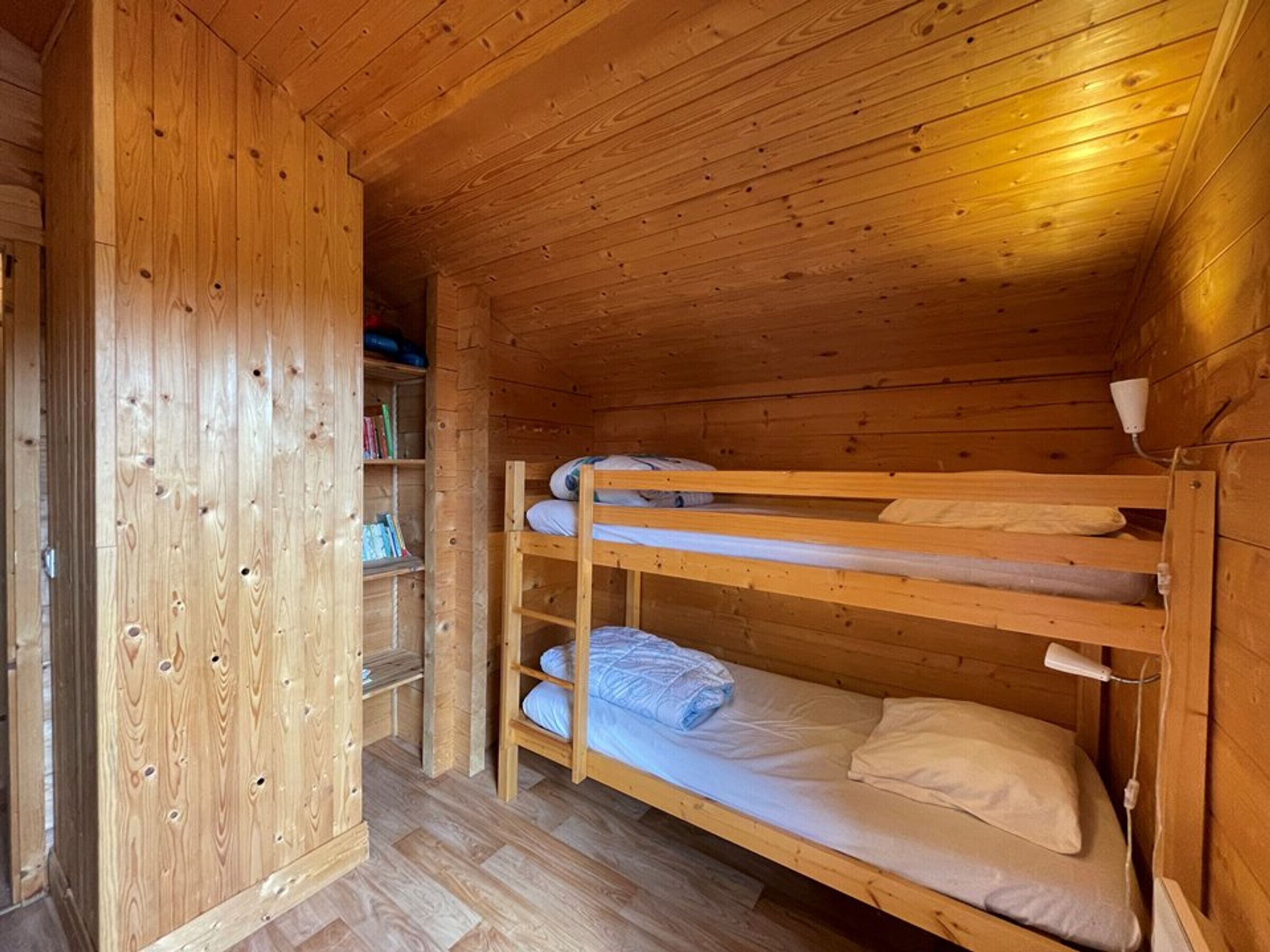 A wooden cabin room with bunk beds, a wooden wardrobe, and a shelf with books.