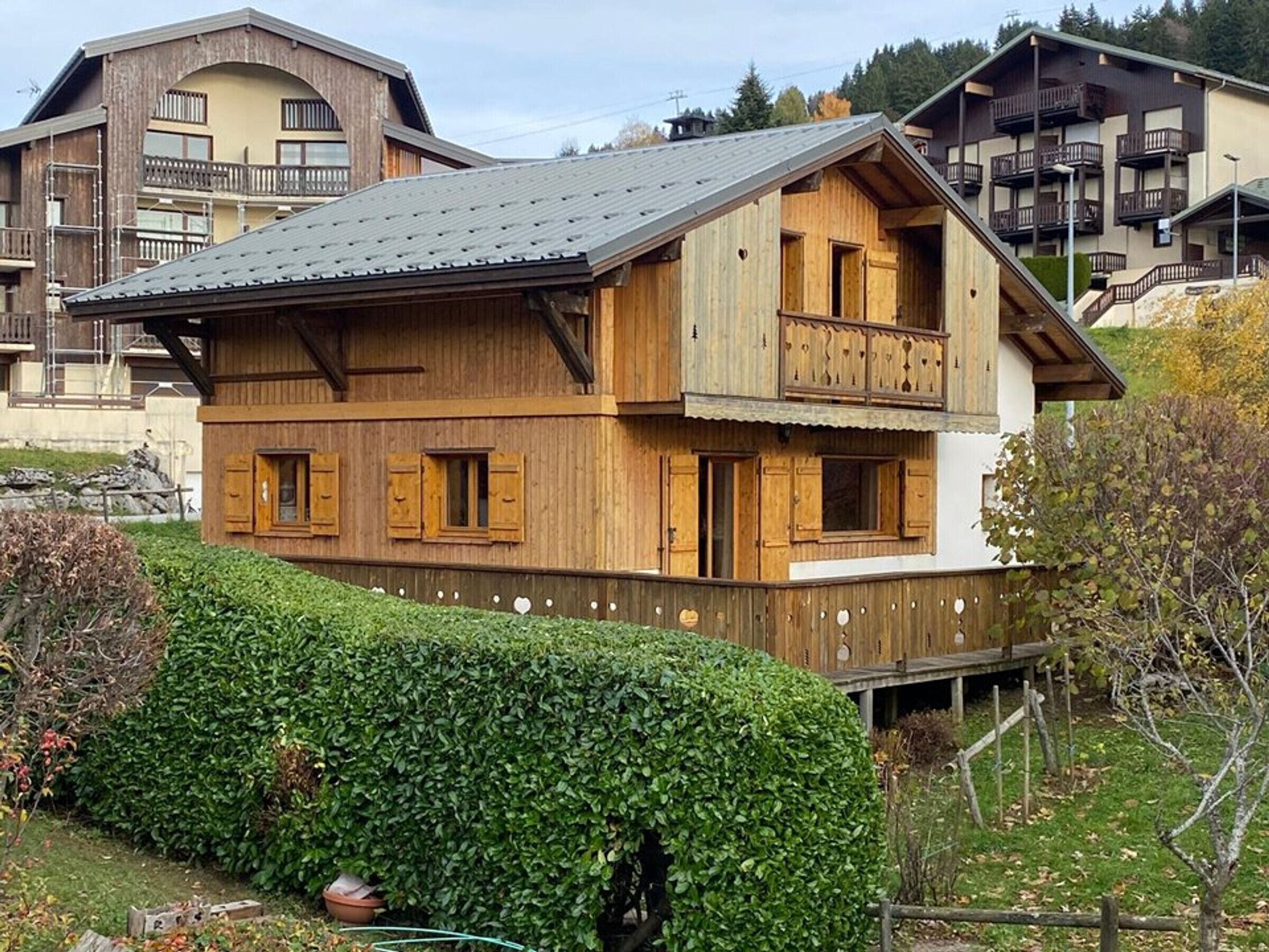 A two-story wooden chalet with a balcony, surrounded by well-maintained hedges and a mountainous backdrop.