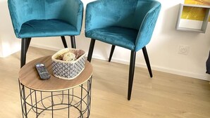 Zwei türkisfarbene Stühle mit schwarzen Beinen, ein kleiner runder Tisch mit einem gewebten Korb und einer Fernbedienung, sowie ein abstraktes gelbes Gemälde an der Wand.
