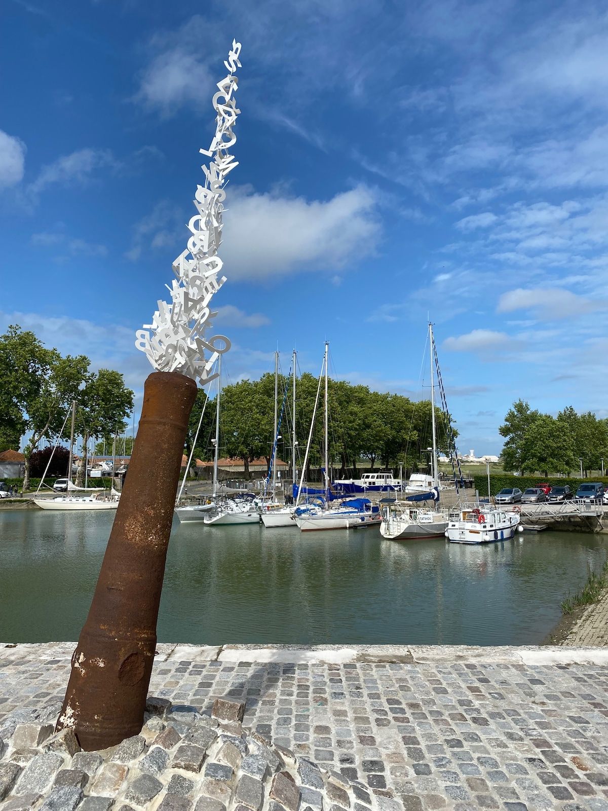 Un muelle con una escultura de metal oxidado, barcos amarrados y un camino empedrado.